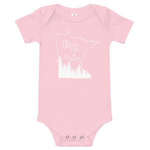 May include: A light pink baby bodysuit with short sleeves. The front features a white outline of the state of Minnesota with the words "Oh Fer Cute" and a forest silhouette inside the state outline.
