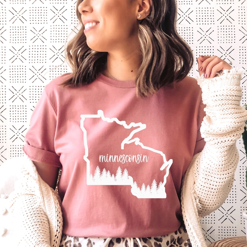 May include: A dusty rose t-shirt featuring a white outline of the state of Minnesota with the word "minnesconsin" in a cursive font. The bottom of the state outline has a white forest design.