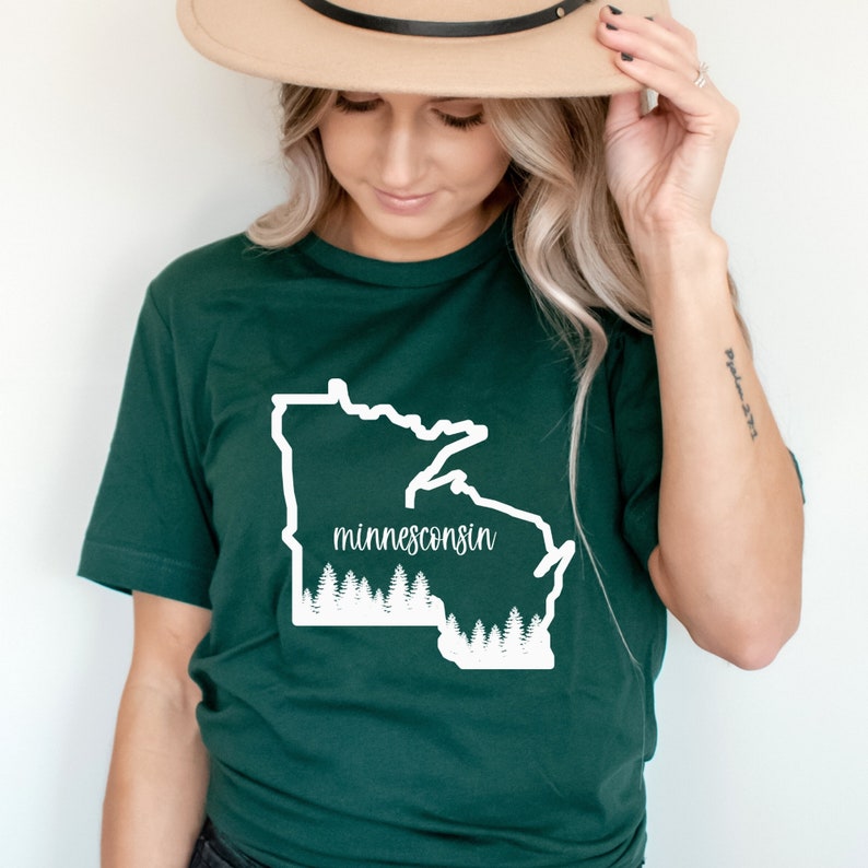 May include: A dark green t-shirt featuring a white outline of the state of Minnesota with the word "minnesconsin" in a script font. Below the state outline is a white graphic of pine trees.