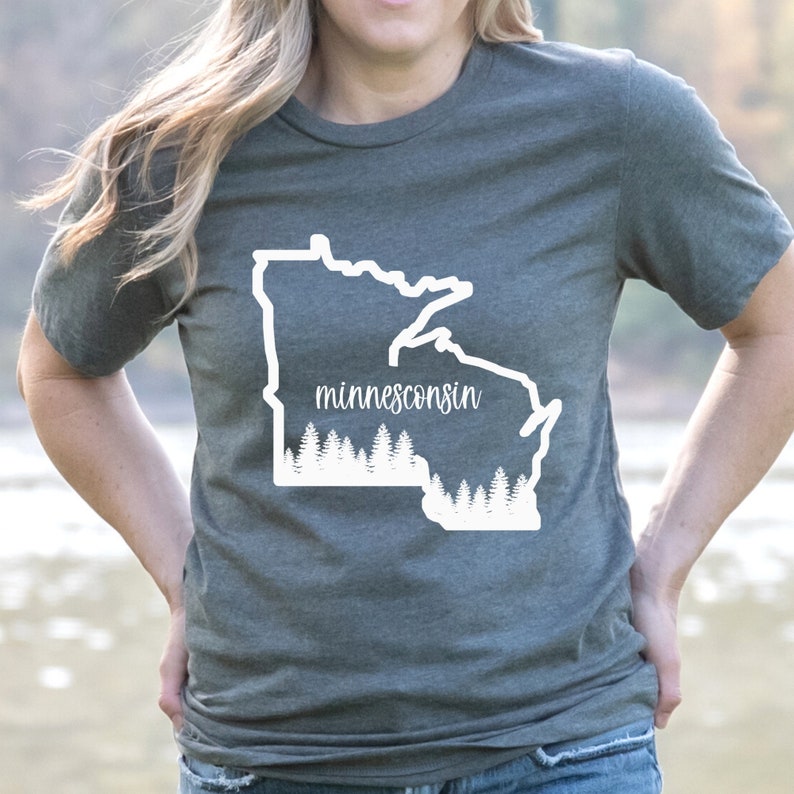 May include: A gray t-shirt featuring a white outline of the state of Minnesota with the word "minnesconsin" in a script font. Below the state outline is a white graphic of a forest.