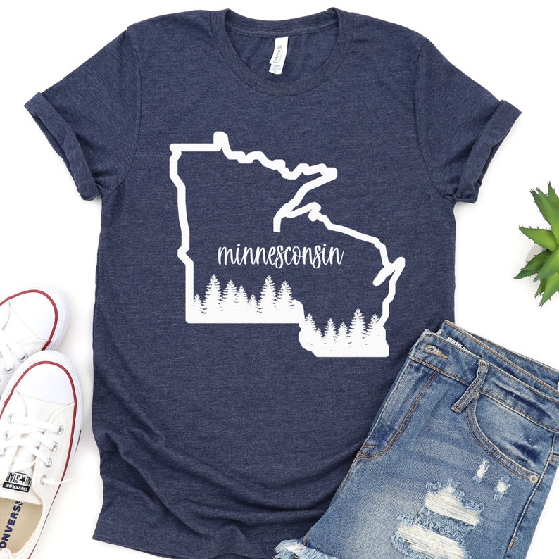 May include: Navy blue t-shirt featuring a white outline of the state of Minnesota with the word "minnesconsin" in a script font. A row of white trees is at the bottom of the state outline. The shirt is folded, with white sneakers and denim shorts visible.