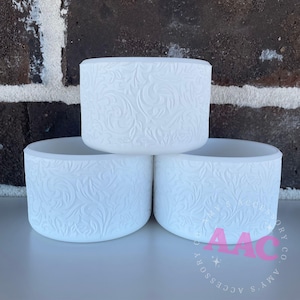 May include: Three white ceramic candle holders with a raised floral design. The holders are stacked on top of each other.