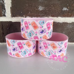 May include: Three pink drink can holders with a repeating pattern of colorful soda cans and flowers. The holders are stacked, showcasing the design. The interior of the holders is a solid pink color.