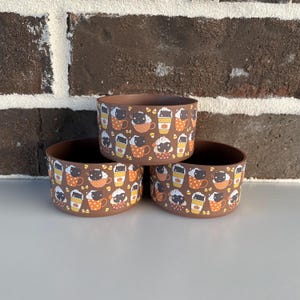 May include: Three brown ceramic bowls stacked on top of each other. Each bowl is decorated with a repeating pattern of coffee cups, pumpkins, and cat faces. The bowls are sitting on a white surface in front of a brick wall.