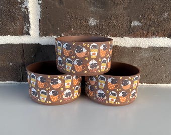May include: Three brown ceramic bowls stacked on top of each other. Each bowl is decorated with a repeating pattern of coffee cups, pumpkins, and cat faces. The bowls are sitting on a white surface in front of a brick wall.