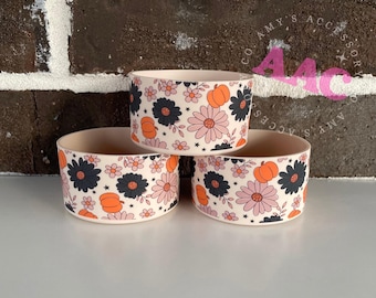 May include: Three beige drink sleeves stacked on top of each other. Each sleeve is decorated with a repeating pattern of black and pink flowers, orange pumpkins, and small white flowers. The sleeves are on a white surface.