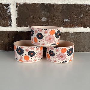 May include: Three stacked ceramic bowls with a floral and pumpkin pattern. The bowls have a cream-colored base and are decorated with black, pink, and orange flowers and pumpkins. The bowls are stacked on top of each other.