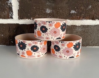 May include: Three stacked ceramic bowls with a floral and pumpkin pattern. The bowls have a cream-colored base and are decorated with black, pink, and orange flowers and pumpkins. The bowls are stacked on top of each other.