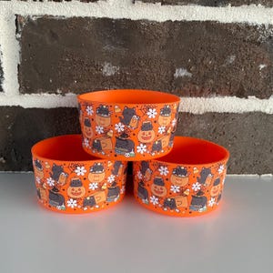 May include: Three orange plastic bowls with a Halloween-themed pattern. The pattern includes black cats, jack-o'-lanterns, spiderwebs, and flowers. The bowls are stacked on top of each other.
