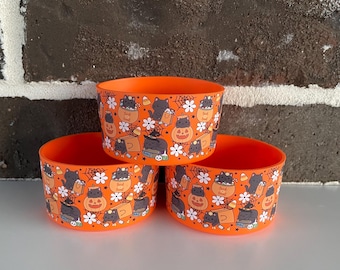 May include: Three orange plastic bowls with a Halloween pattern. The pattern features black cats, jack-o'-lanterns, spiderwebs, and flowers. The bowls are stacked together.