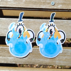 May include: Two holographic stickers featuring a cartoon character with a top hat, one eye, and a bow tie. The character is primarily blue with yellow and black accents. The stickers are on a wooden surface.