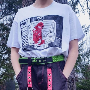 Op de afbeelding: Wit T-shirt met een grafische print van een rode figuur op een computerscherm, met het woord "Welcome" en de letters "NT". De outfit omvat een zwarte broek, een groene riem met studs en roze bretels.
