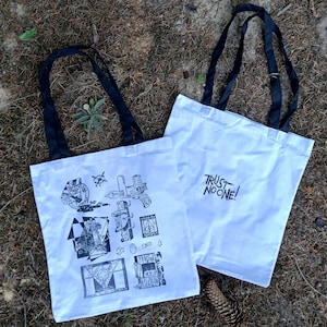 May include: Two white tote bags with black handles rest on a bed of brown leaves and pine needles. One bag features a black and white illustration, the other reads "TRUST NO ONE!" in black lettering. The bags are made of a sturdy fabric.