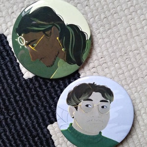 May include: Two round button badges with illustrations of people. The first badge features a person with long dark green hair, wearing glasses and a green shirt. The second badge features a person with short dark hair, wearing glasses and a green shirt.
