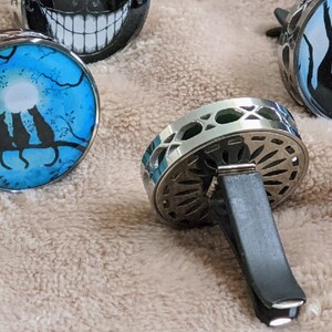 May include: Three silver car air fresheners with different designs. One features a moon and three black cats on a branch, another has a black and white silhouette of a tree and a cat, and the third is a silver and black decorative design.