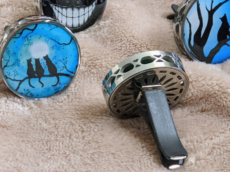 May include: Three silver car air fresheners with different designs. One features a moon and three black cats on a branch, another has a black and white silhouette of a tree and a cat, and the third is a silver and black decorative design.