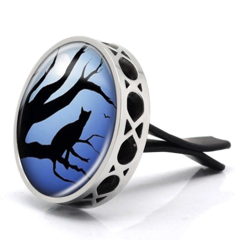 May include: A silver car air freshener featuring a black cat silhouette perched on a tree branch against a blue moonlit sky. The air freshener has a round glass insert and a metal frame.