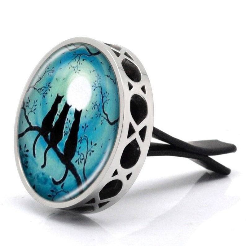 May include: A silver car air freshener with a round glass insert featuring a blue and white moonlit scene with three black cats silhouetted on a branch.