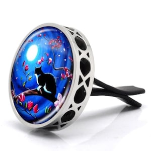 May include: A silver car air freshener featuring a black cat perched on a branch beneath a full moon. The cat is black and white with pink blossoms in the background.