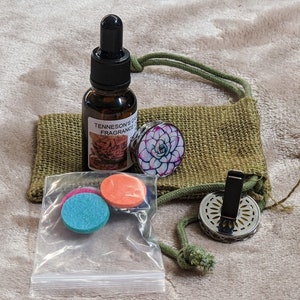 May include: A set of car air freshener supplies including a small bottle of essential oil, two felt pads, a metal car vent clip, and a decorative round disc with a floral design. The items are packaged in a small hessian bag with a drawstring.