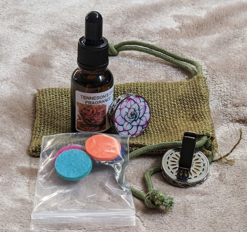 May include: A set of car air freshener supplies including a small bottle of essential oil, two felt pads, a metal car vent clip, and a decorative round disc with a floral design. The items are packaged in a small hessian bag with a drawstring.