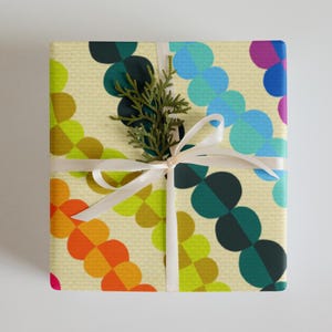 May include: A gift wrapped in colourful wrapping paper with a geometric pattern of overlapping circles in shades of green, blue, yellow, orange, red, and purple. The paper is cream coloured with a subtle texture. A white ribbon is tied around the box with a sprig of greenery on top.