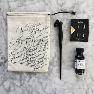 May include: A white canvas bag with black drawstring, featuring a handwritten quote about calligraphy and the power of the written word. The bag is accompanied by a black calligraphy pen, a bottle of black ink, and a card with calligraphy nibs.