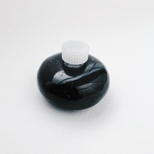 Puede incluir: Una botella redonda de cerámica negra con una tapa de plástico blanco. La botella tiene un acabado liso y brillante.
