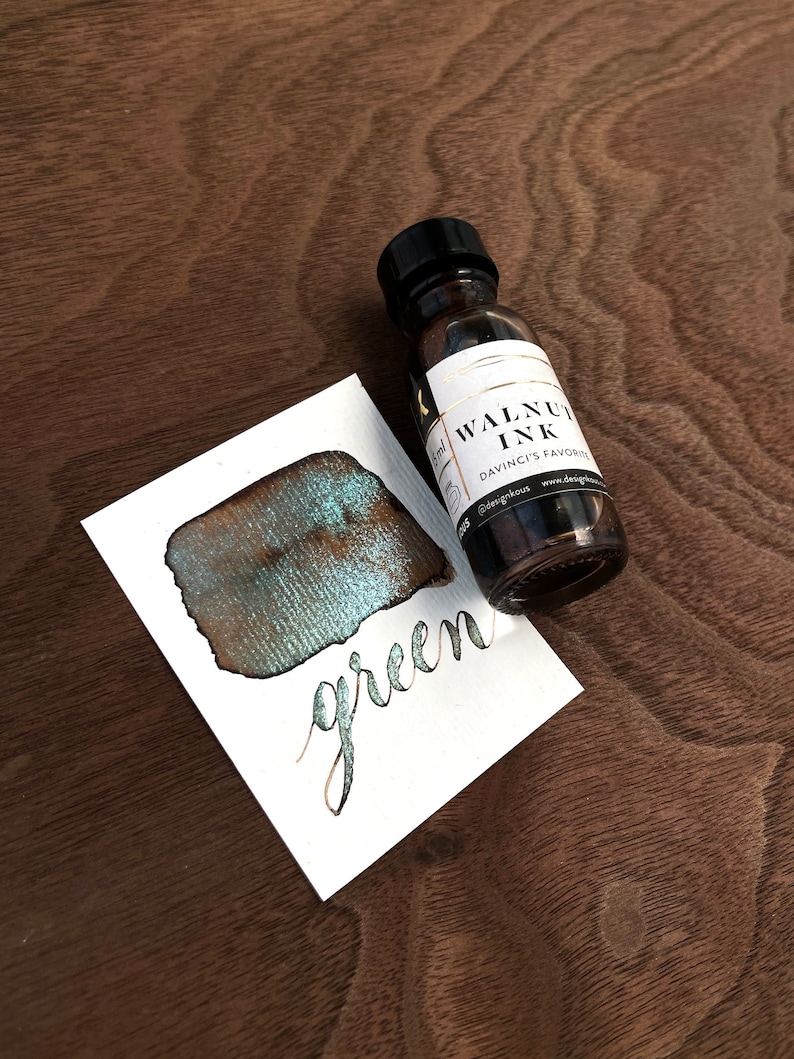 May include: A bottle of Walnut Ink by Davinci's Favorite sits next to a white card with a green ink swatch and the word "green" written in cursive. The ink bottle has a black cap and a white label with gold trim.