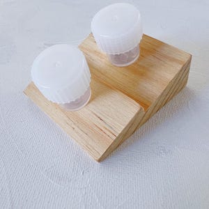 Pode incluir: Dois estojos de plástico branco para lentes de contato em um suporte de madeira. O suporte é inclinado e feito de madeira marrom claro.