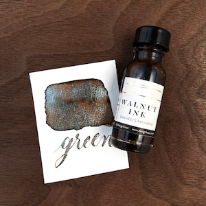 May include: A bottle of walnut ink next to a swatch of ink on a white card. The ink swatch is a dark brown color with green and gold sheen. The card has the word "green" written in cursive. The ink bottle has a black cap and a white label.