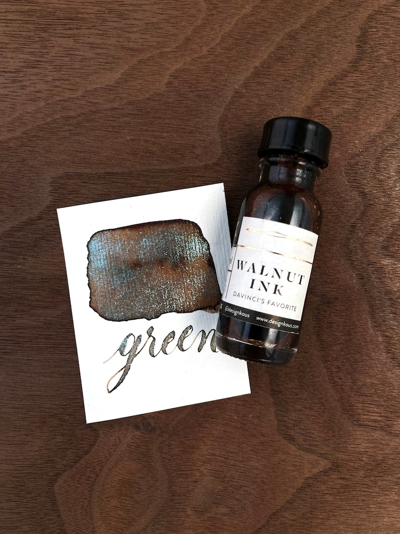 May include: A bottle of walnut ink next to a swatch of ink on a white card. The ink swatch is a dark brown color with green and gold sheen. The card has the word "green" written in cursive. The ink bottle has a black cap and a white label.