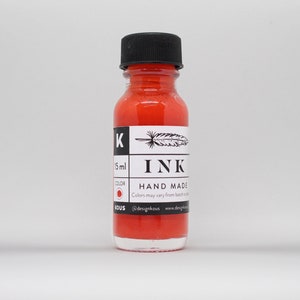 Puede incluir: Una pequeña botella de vidrio de tinta roja etiquetada "INK HAND MADE" con una ilustración de pluma y pluma. La botella es de 15 ml y el color es rojo. La etiqueta también incluye el texto "@designkous www.designkous.com".