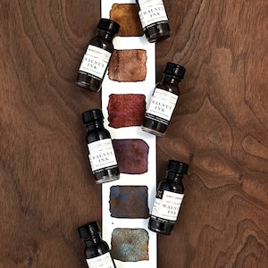 May include: An overhead shot displays six small bottles of walnut ink with black caps and white labels. The labels read "WALNUT INK" and "Davinci's Favorite." Swatches of ink in various shades of brown and blue-green are displayed on a white card.
