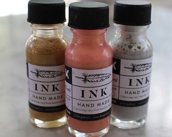 Puede incluir: Tres pequeños frascos de vidrio de tinta, cada uno con una tapa negra y una etiqueta blanca. Las etiquetas presentan un diseño de pluma y la palabra "INK" en letras negras. Los colores de la tinta son marrón, rosa y gris.