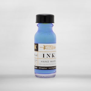 May include: A small glass ink bottle filled with light blue ink, featuring a black cap. The label reads "INK HAND MADE" with a gold feather illustration. The bottle contains 5ml of ink.