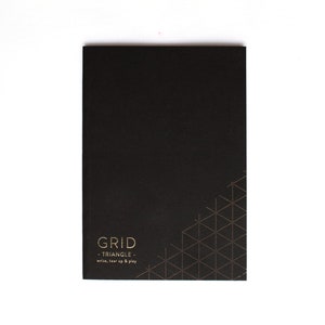 May include: A black notebook with the text "GRID - TRIANGLE - write, tear up & play" in gold. A gold triangle pattern is in the lower right corner. The notebook is rectangular and has a matte finish.