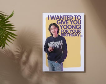 Op de afbeelding: Een poster met de tekst "I WANTED TO GIVE YOU YOONGI FOR YOUR BIRTHDAY..." toont een persoon in een marineblauw sweatshirt en een blauwe jeans. De achtergrond is geel met een beige rand. De poster hangt aan een beige muur.