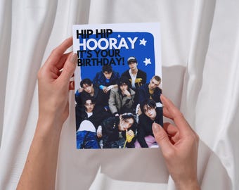 May include: A birthday card held by hands, featuring a group of people. The card has the words "HIP HIP HOORAY IT'S YOUR BIRTHDAY!" in black and white text on a blue background with stars.