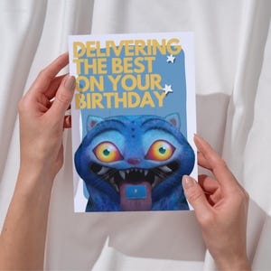 May include: A birthday card with the text "DELIVERING THE BEST ON YOUR BIRTHDAY" in yellow. The card features a blue cartoon cat with large yellow eyes and a tongue sticking out. Two small white stars are also on the card.