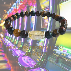 K&ouml;nnte beinhalten: Ein dreifach sch&uuml;tzendes Gl&uuml;cksarmband mit schwarzen, braunen und gr&uuml;nen Perlen und einer klaren Quarzperle mit silbernen Akzenten. Das Armband ist vor einer Reihe von Spielautomaten fotografiert.
