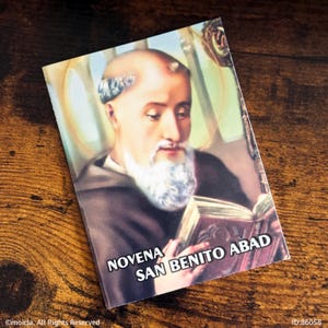 May include: A rectangular card featuring an illustration of a man with a white beard, reading a book. The card has the text "NOVENA SAN BENITO ABAD" in white. The image has a warm color palette and is set against a wooden background.