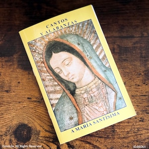 May include: A yellow book titled "CANTOS Y ALABANZAS" with an illustration of a religious figure. The image features a woman with a blue veil and a halo. The text "A MARÍA SANTÍSIMA" is also visible.