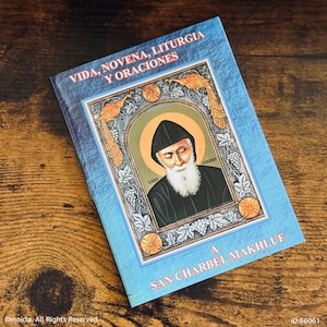 May include: A book with a blue cover featuring an illustration of a saint with a white beard and black attire. The book's title includes the words "Vida, Novena, Liturgia Y Oraciones" and "San Charbel Makhluf". The image is on a wooden surface.