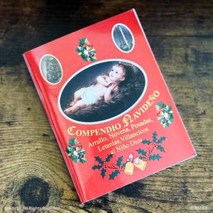 May include: A red book with the title "COMPENDIO NAVIDEÑO" and an image of baby Jesus. The cover features decorative elements like holly, bells, and floral arrangements. The book's title is in a decorative font.
