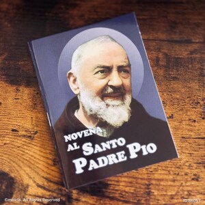 May include: A rectangular booklet featuring a portrait of Padre Pio with a white beard and a halo. The text "NOVENA AL SANTO PADRE PIO" is printed in white on a dark background. The booklet is on a wooden surface.