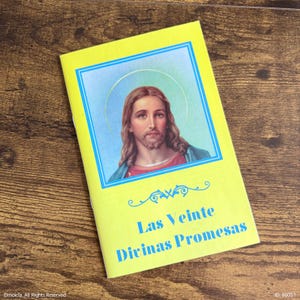 May include: A yellow booklet with a blue border and the image of Jesus Christ. The text "Las Veinte Divinas Promesas" is printed in blue. The booklet is lying on a wooden surface.