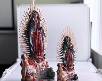May include: Two resin statues of the Virgin Mary of Guadalupe, one 12 inches tall and the other 8 inches tall. Both statues feature the Virgin Mary in a blue and red robe with a golden halo, standing on a base with a cactus and two figures.