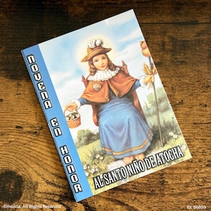 May include: A religious booklet with a blue spine and a colorful illustration. The cover features a depiction of a young figure in a blue dress, brown cape, and hat, holding a basket and staff. The text on the cover reads "NOVENA EN HONOR AL SANTO NIÑO DE ATOCHA."