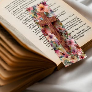 May include: A bookmark in the shape of a wooden cross, adorned with a floral pattern of pink, purple, and yellow blooms. It rests within an open book, its pages showing a vintage, aged appearance.